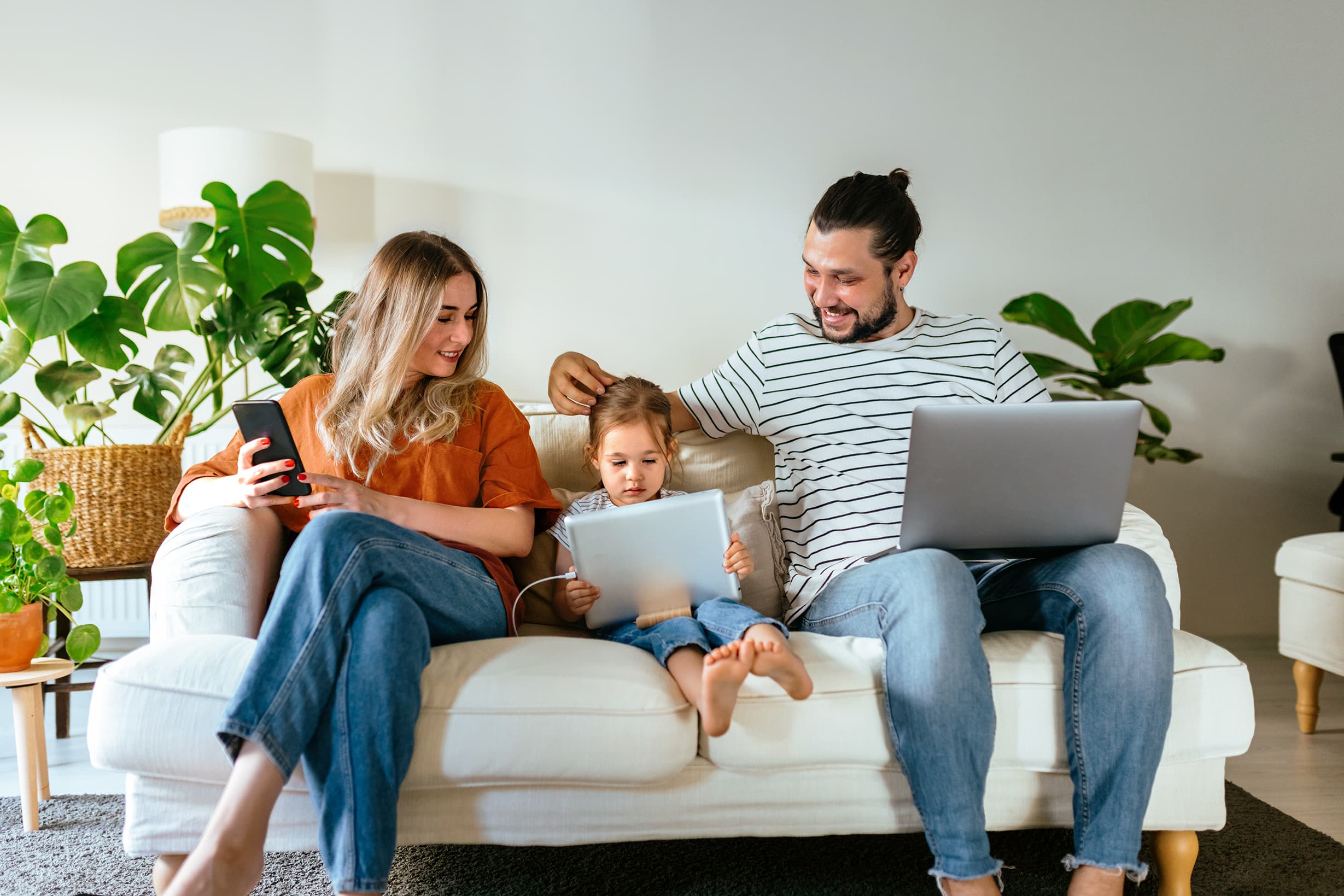 Happy family of three sitting on a white couch at home, parents with laptop and phone while young daughter draws on paper.